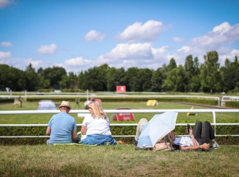 Rennbahnbesucher sitzen in der langen Pause am verlassenen Führring in der Sonne